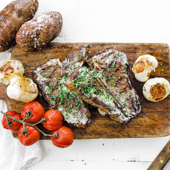 grilled steaks on a cutting board with roasted garlic, tomatoes and potatoes