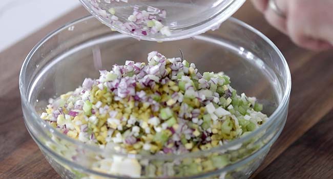 adding chopped ingredients to a bowl of potato salad