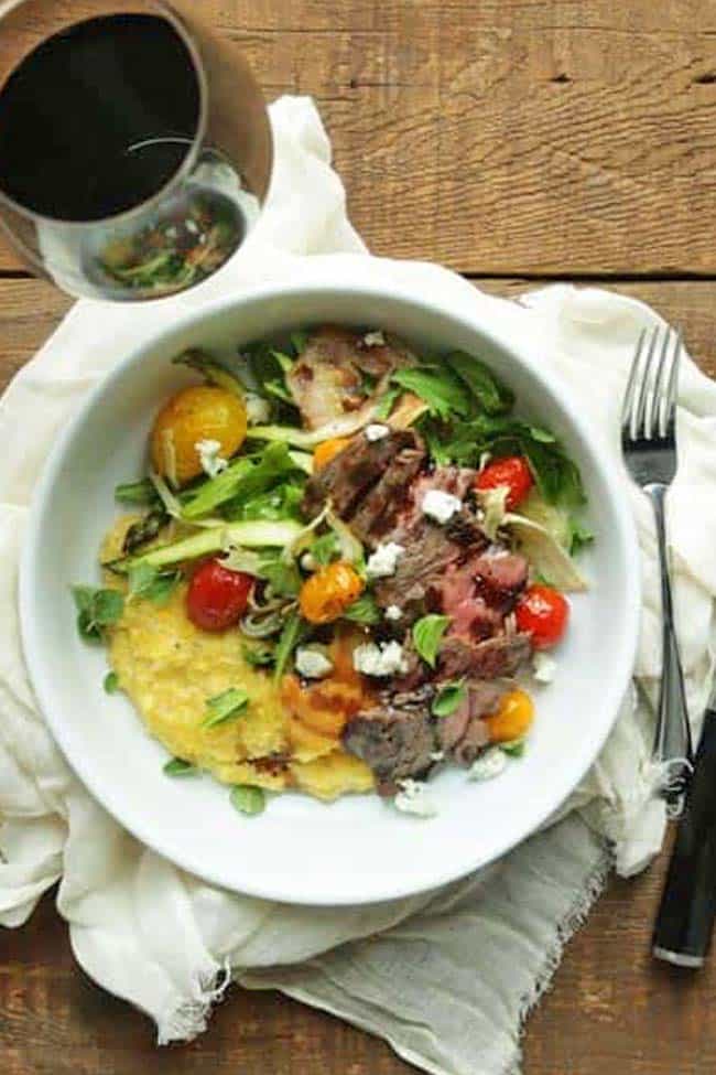 grilled sliced skirt steak with vegetables, herbs and polenta in a bowl with a glass of red wine
