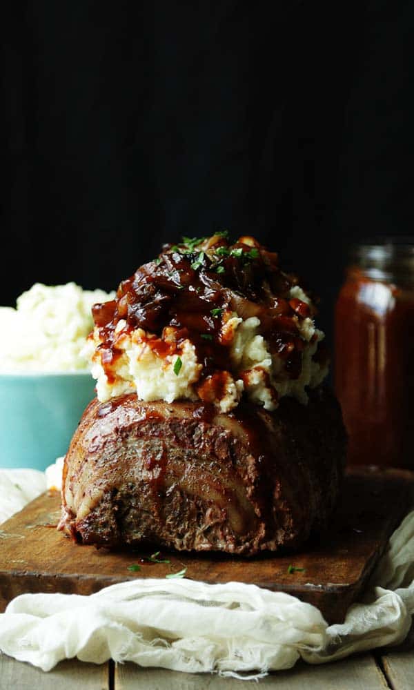 a bacon wrapped meatloaf that has mashed potatoes, caramelized onions and bbq sauce on top