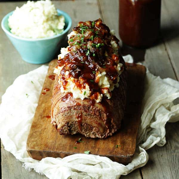 meatloaf wrapped in bacon with mashed potatoes, onions and BBQ sauce on a cutting board