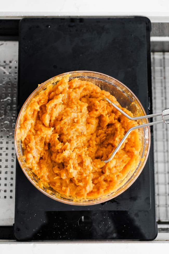 a hand masher mashing baked sweet potatoes in a bowl