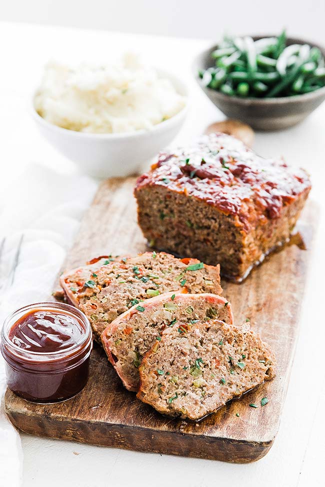 sliced meatloaf on a cutting board next to mashed potatoes and green beans