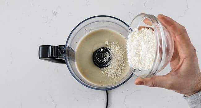 pouring in grated parmesan cheese to caesar salad dressing in a food processor