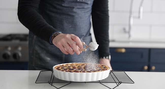 adding powdered sugar to a cooked clafoutis on a cooling rack