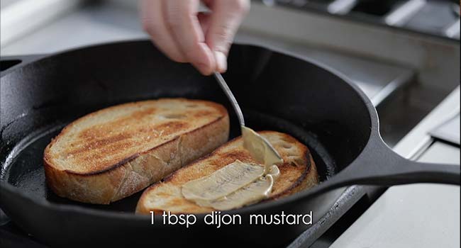 spreading dijon mustard on a toasted slice of bread in a pan