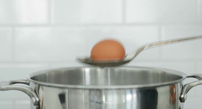 adding an egg on a spoon to a pot of boiling water
