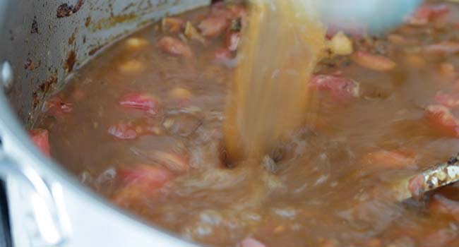 pouring beef stock into a pot of caramelized vegetables and beans