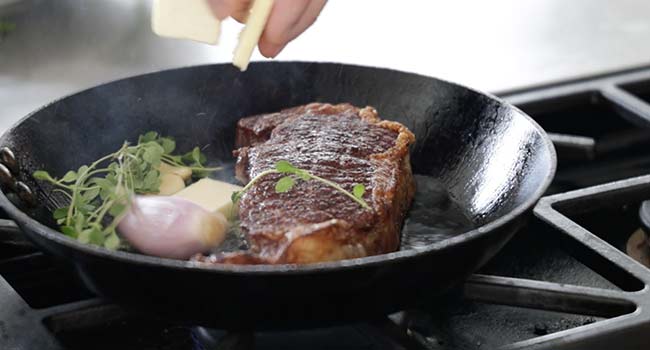 adding herbs and butter to a pan with a steak