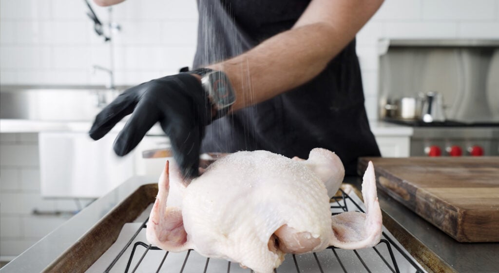 sprinkling salt on a whole chicken on a rack on a sheet tray