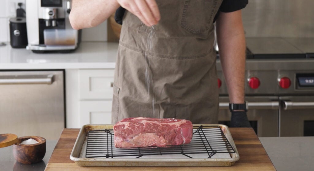 chef sprinkling salt on a piece of beef on a rack over a tray