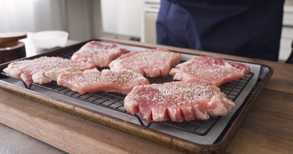Pork on a rack over a sheet tray, seasoned with salt and pepper