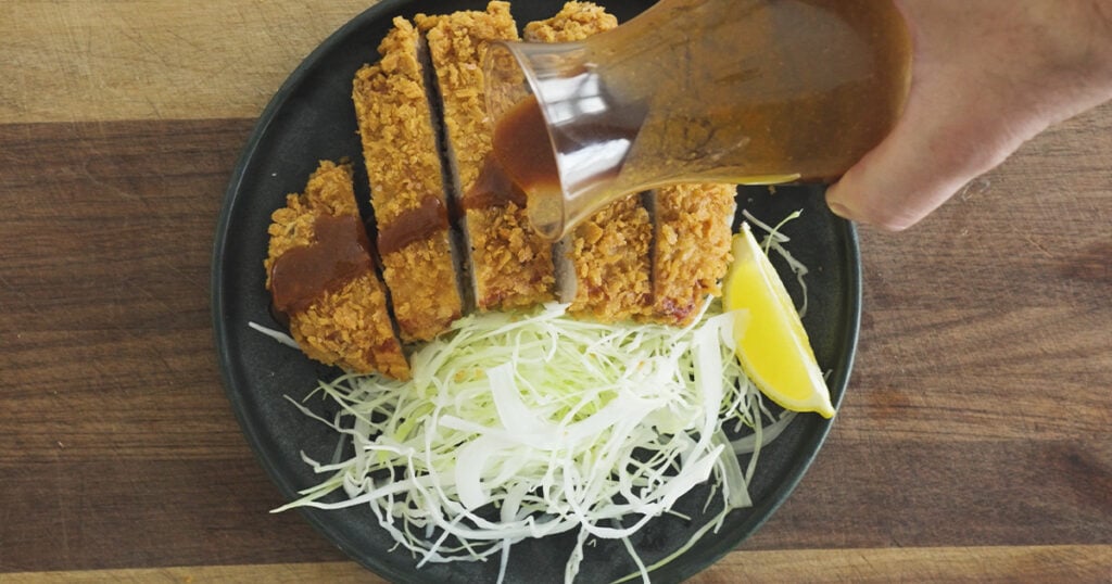 Pouring sauce on a sliced  piece of fried pork on a plate with shredded cabbage and a lemon wedge on the side