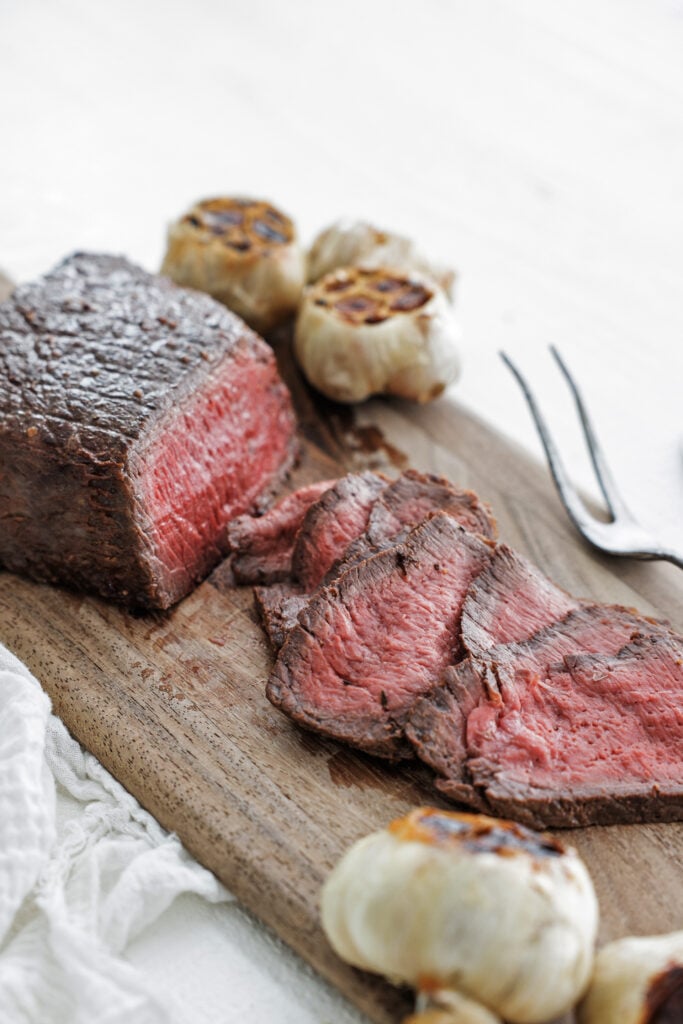London broil with serving fork and roasted garlic on a wooden board