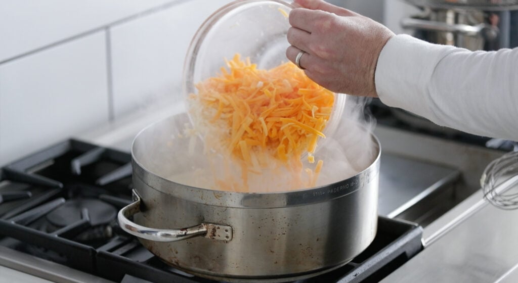 adding shredded cheese to a large pot with milk