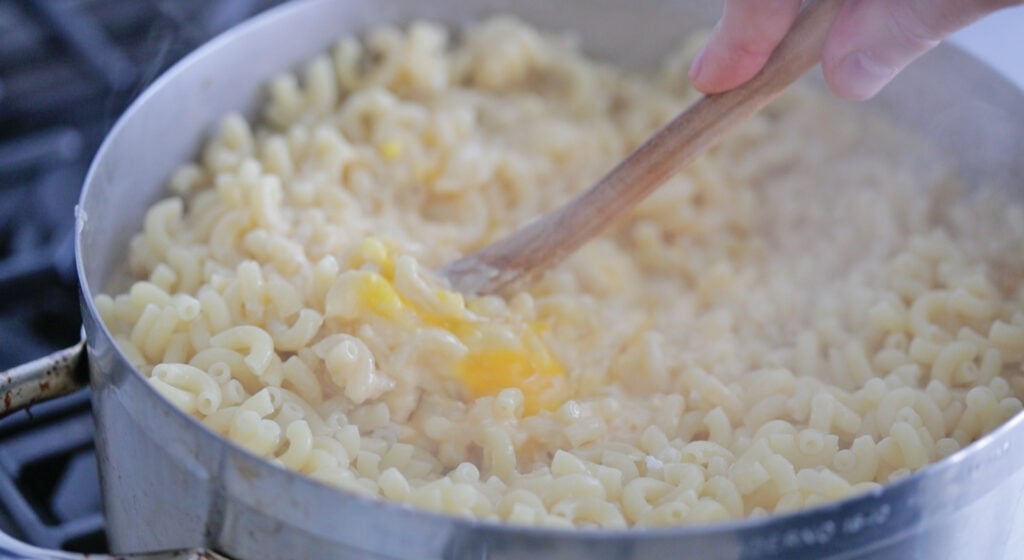 mixing together eggs, cheese sauce and cooked macaroni noodles together in a pot