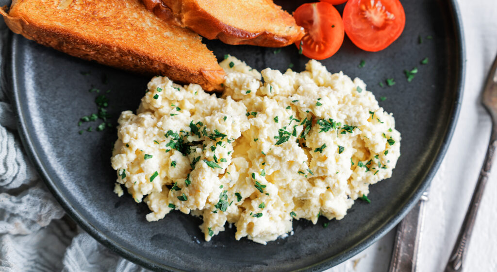 Scrambled eggs topped with parsley on a plate with toast and sliced cherry tomatoes on the side
