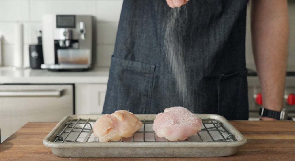 Chicken breasts on a rack over a pan being sprinkled with salt. 