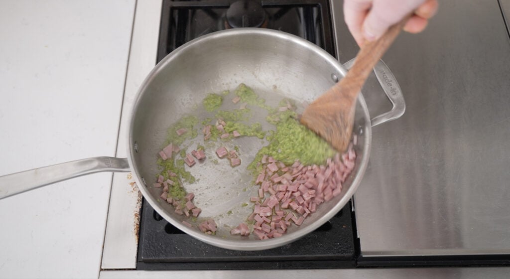 Made in brand pot on a stove with ham and sofrito being mixed with a wooden utensil.