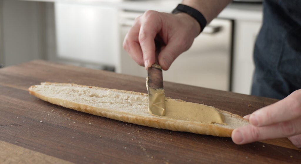 Using a knife to spead mustard on a half of a baguette