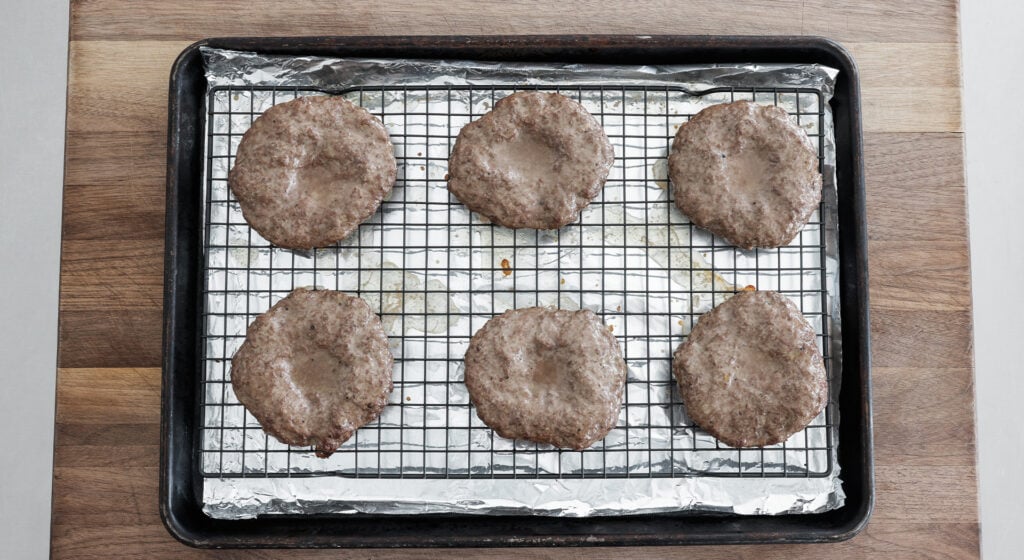 6 browned burger patties on a rack over a sheet tray lined with foil