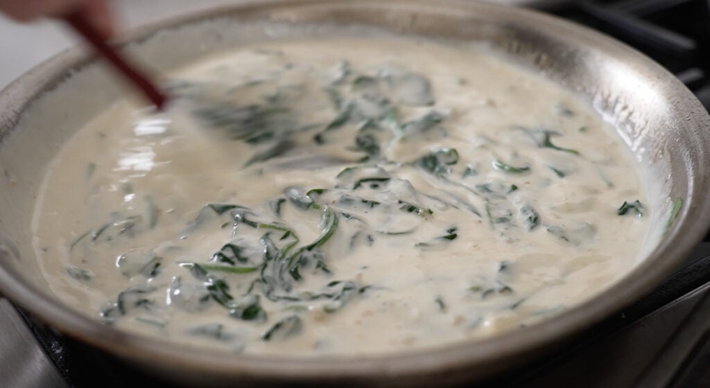 A kitchen utensil stirring sauce and spinach together