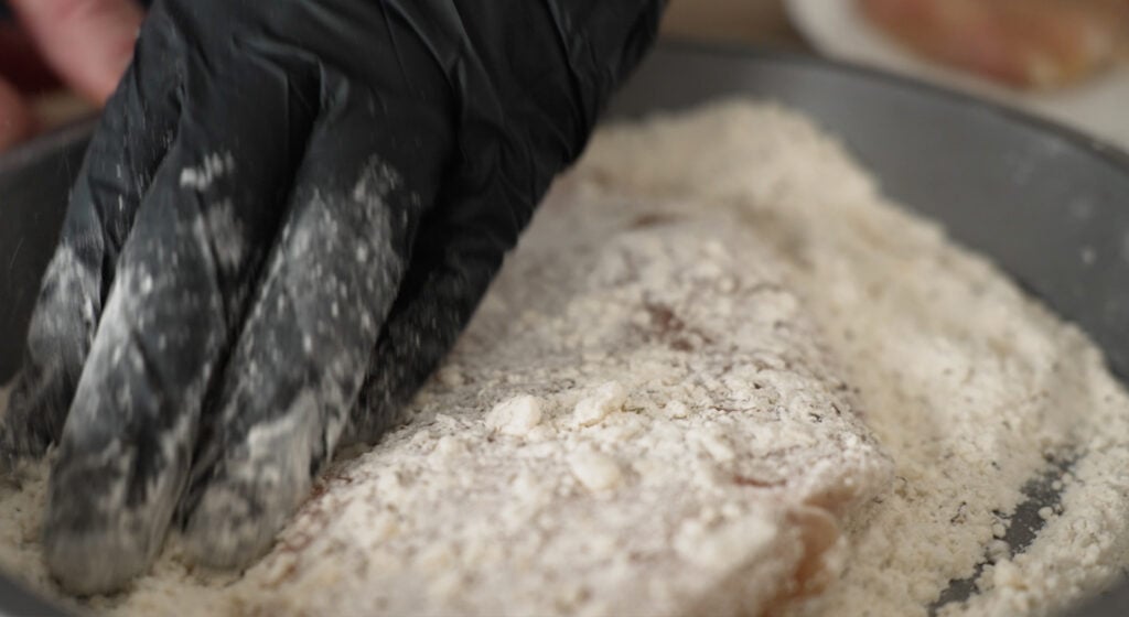 A gloved hand coating a chicken breast in flour