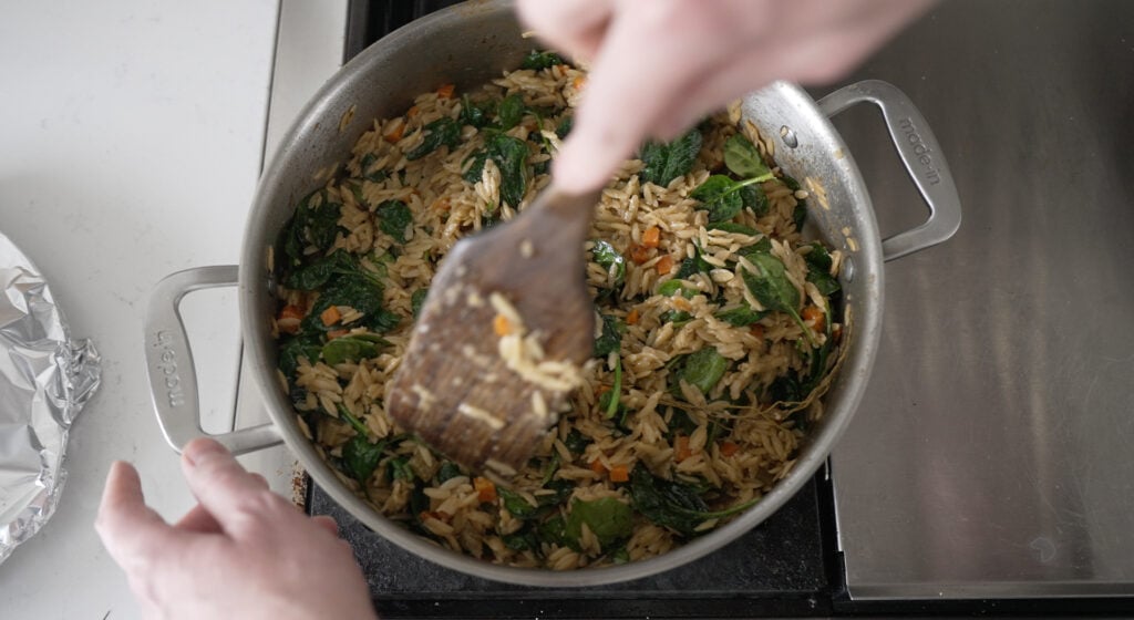 using a wooden utensil to stir spinach into the pot with orzo