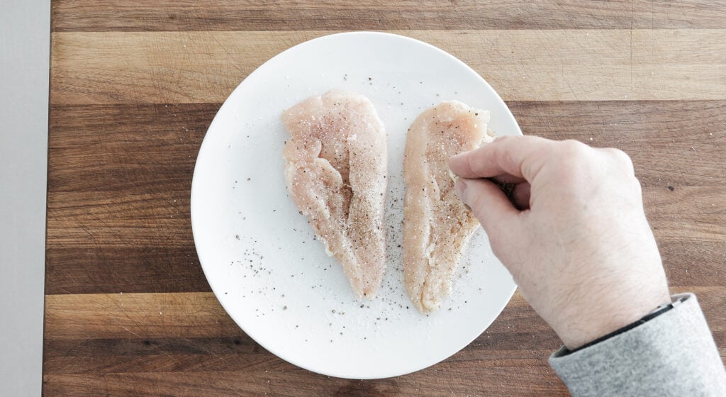 Seasoning two chicken breasts on a white plate
