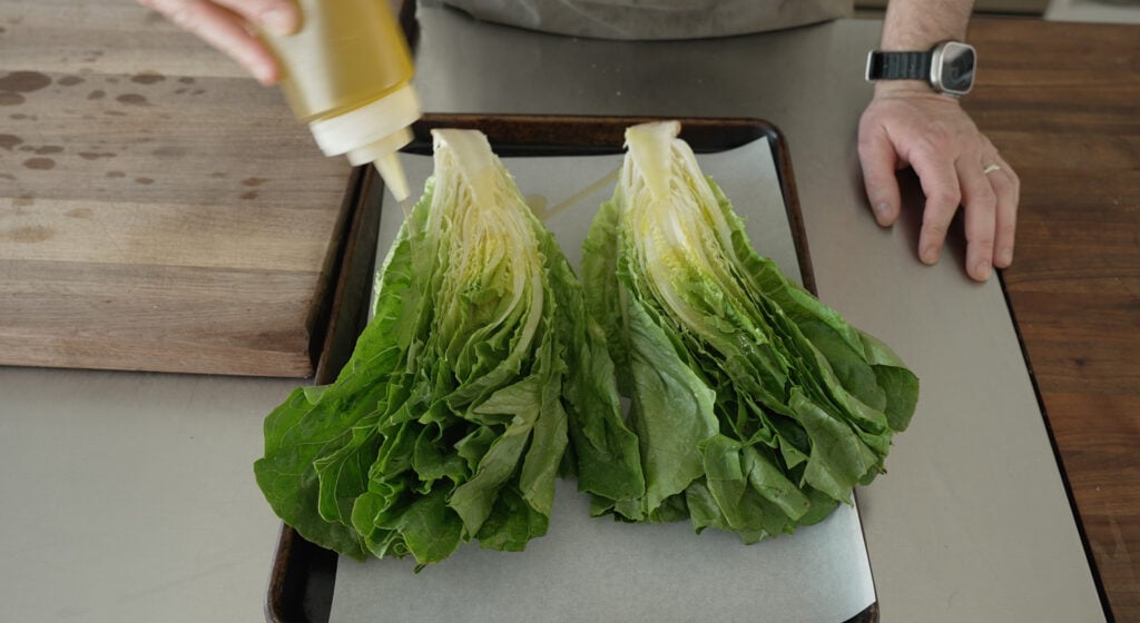 a halved head of romaine on a sheet tray being drizzled with olive oil