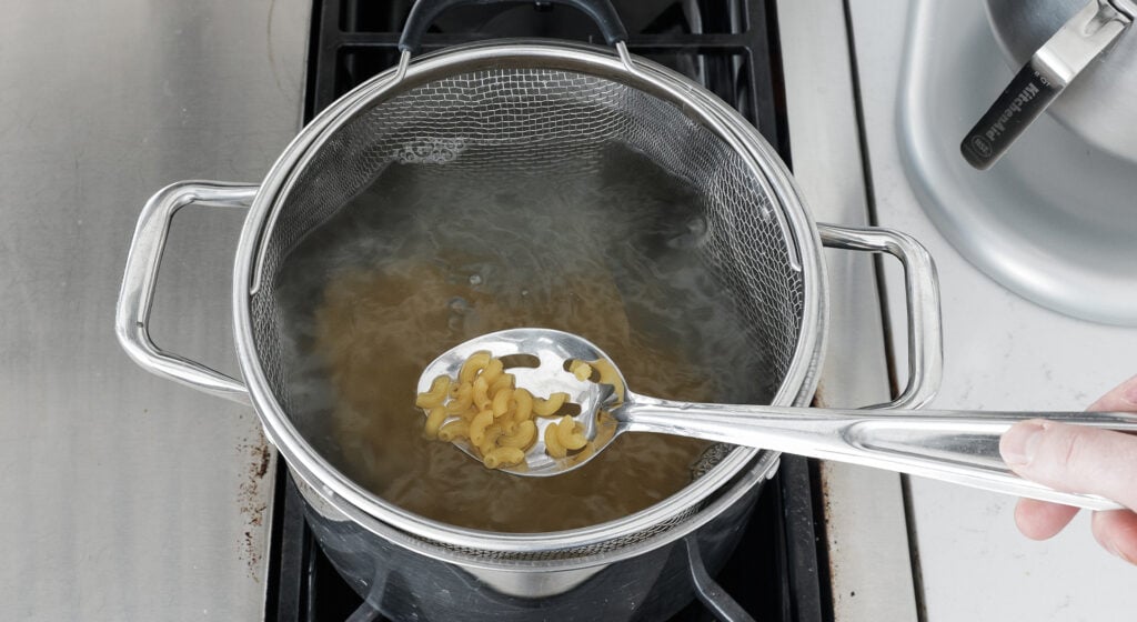 a slotted spoon containing macaroni noodles being held above a pot of water