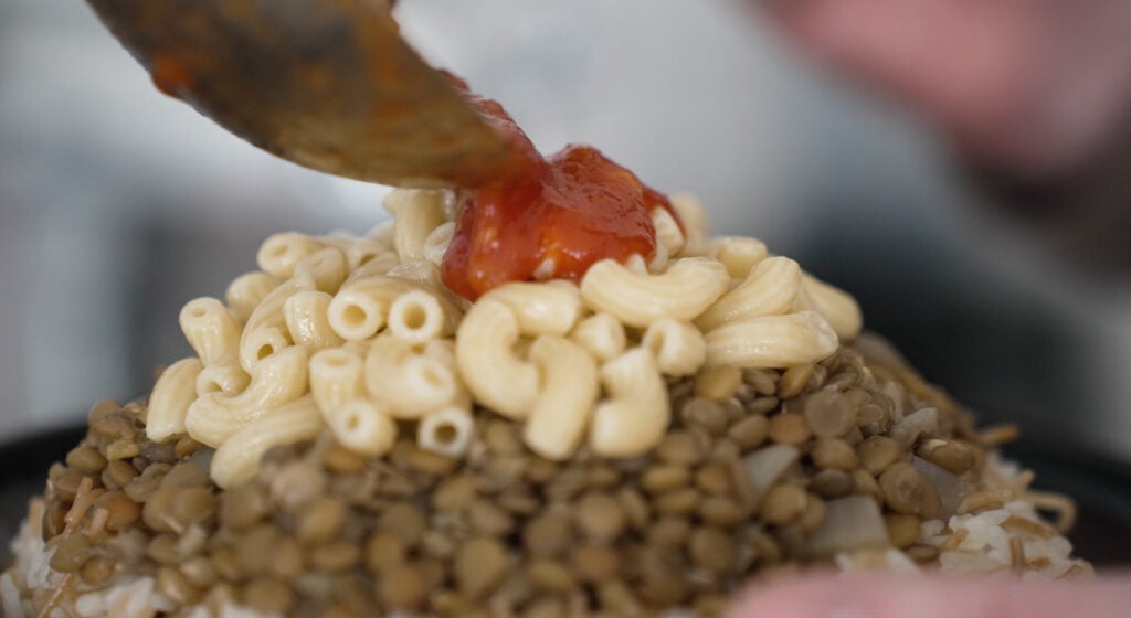 Spooning tomato sauce on top of macaroni noodles, layered on cooked lentils