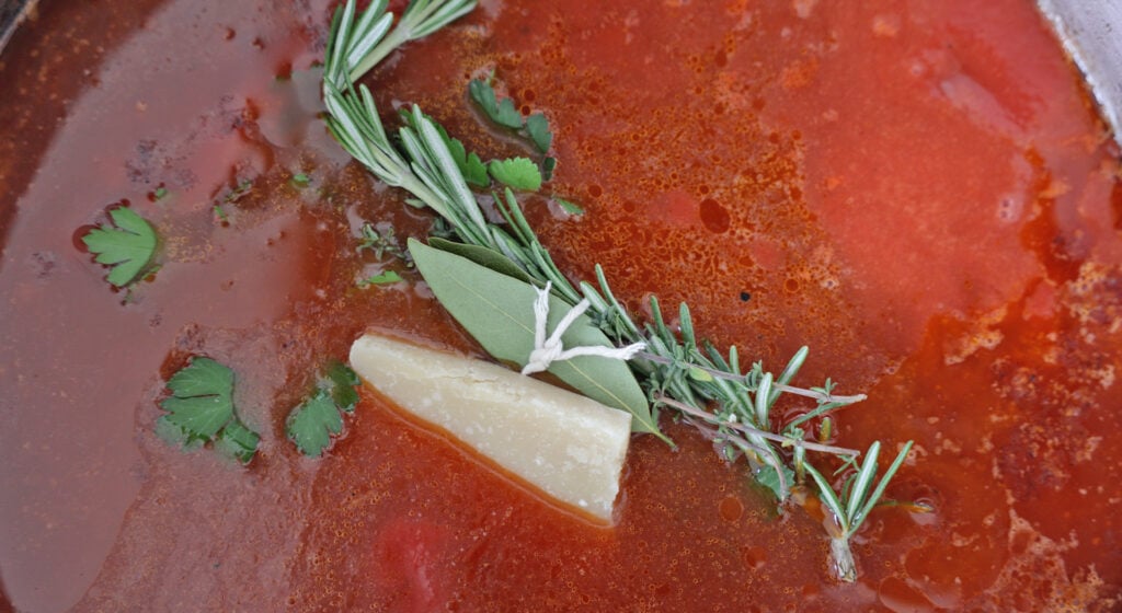 close up of red sauce with fresh herbs and a cheese rind on the top, not yet stirred in