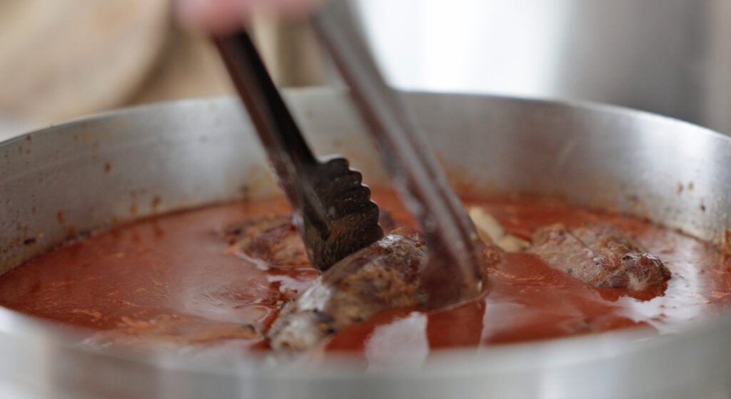 tongs placing pieces of lamb into the pot with the red sauce