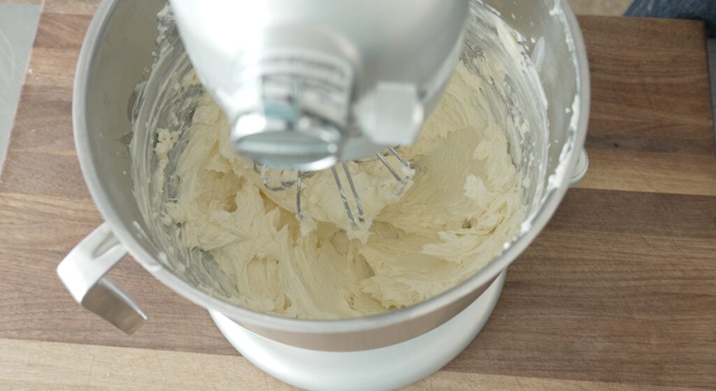 a stand mixer with a whisk attachment and a bowl containing cream cheese and mascarpone