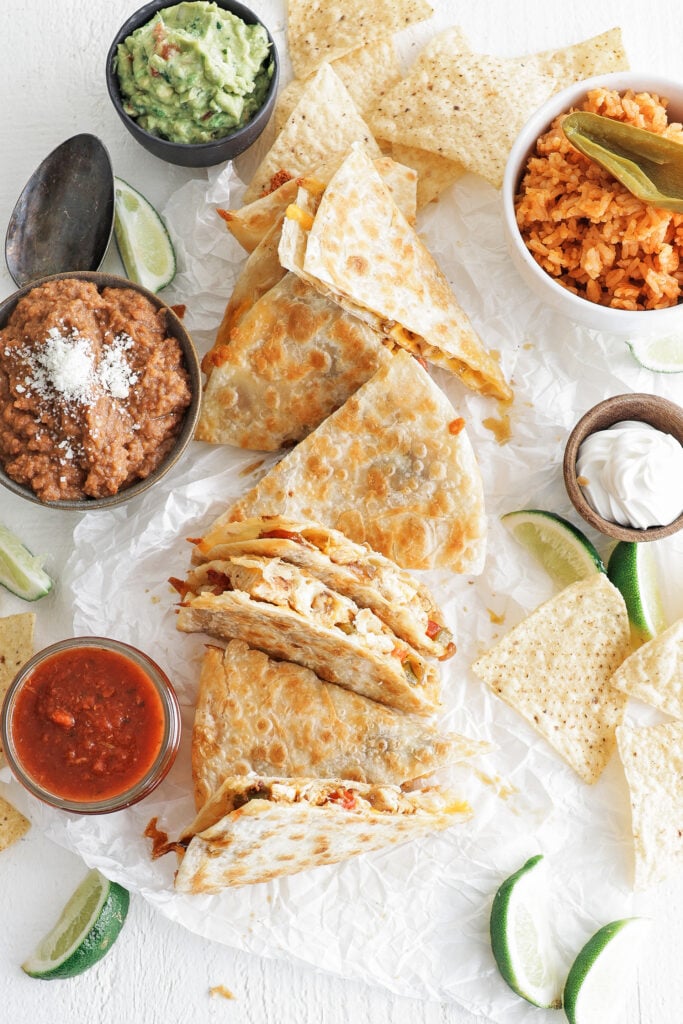 Chicken quesadillas cut into pieces, next to wedges of lime, salsa, sour cream, refried beans, guacamole, corn chips and rice