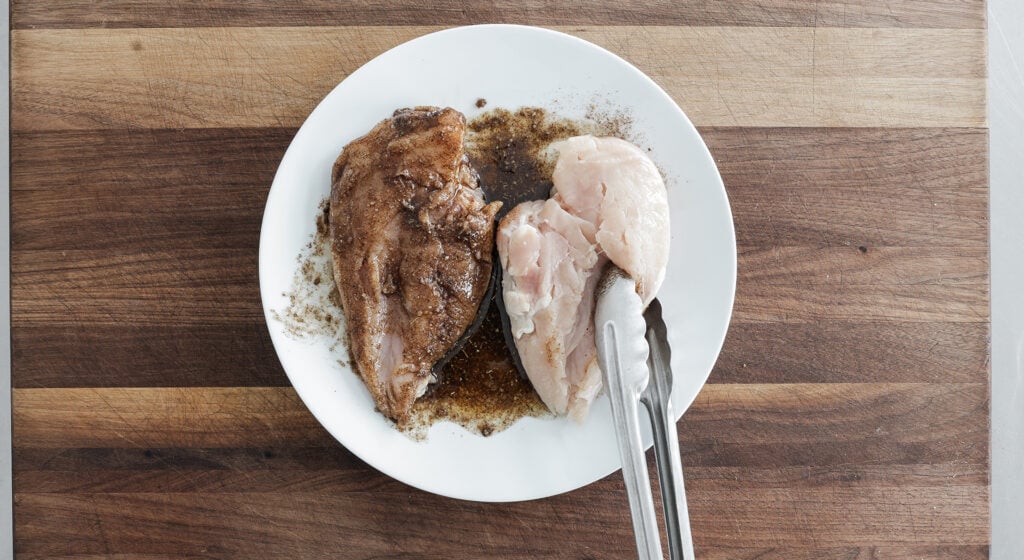 A white plate containing one chicken breast coated in seasoning and one not coated