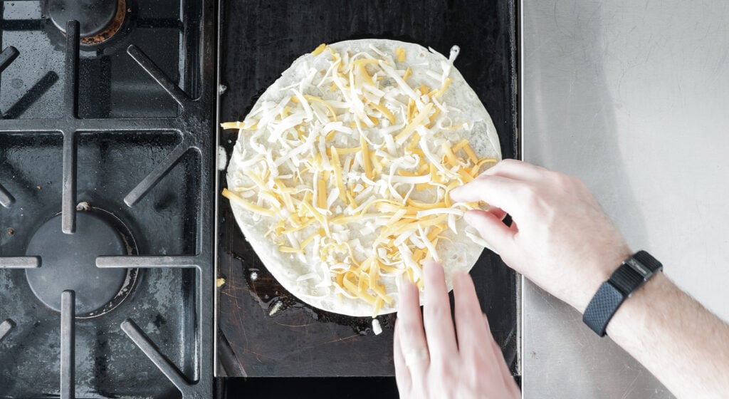 spreading shredded cheese on a flour tortilla on a griddle on the stove