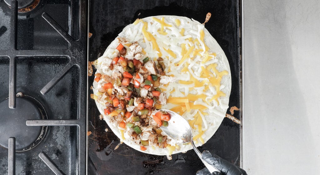 a tortilla on a griddle with shredded cheese on one side and the chicken and veggie mixture on the other half