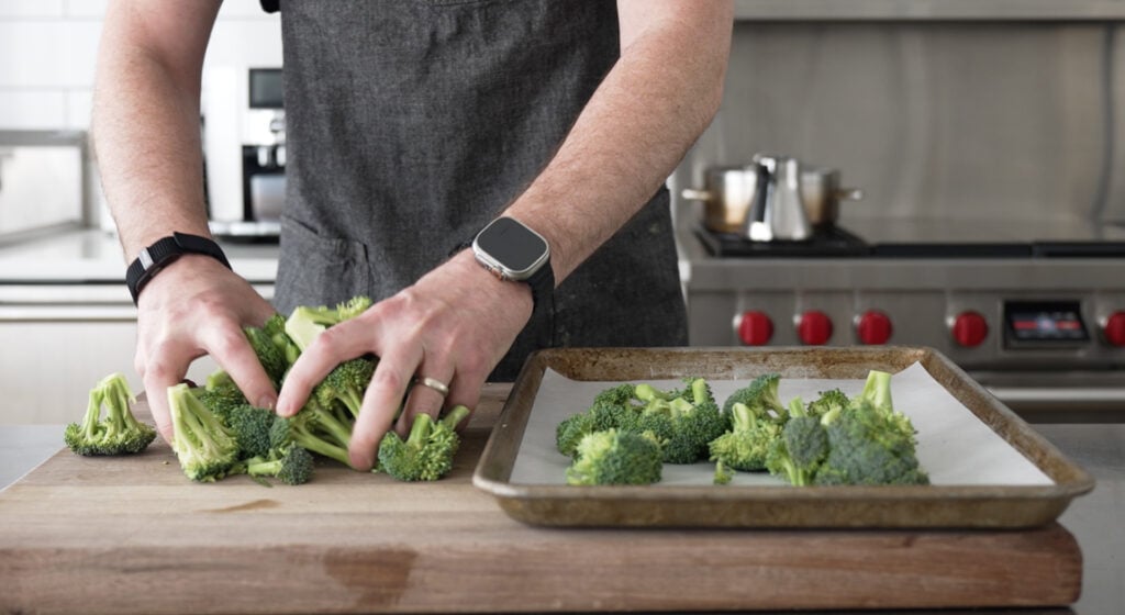 two hands moving broccoli from a cutting board to a sheet tray lined with parchment paper