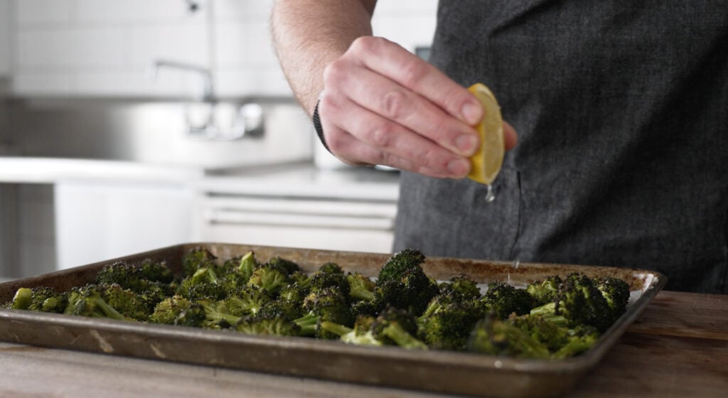 a hand squeezing a half of a lemon over a tray of broccoli