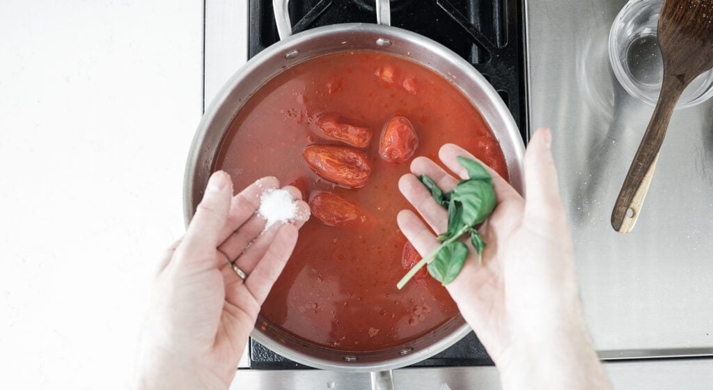 hands holding basil and salt over a pot with tomato-y liquid