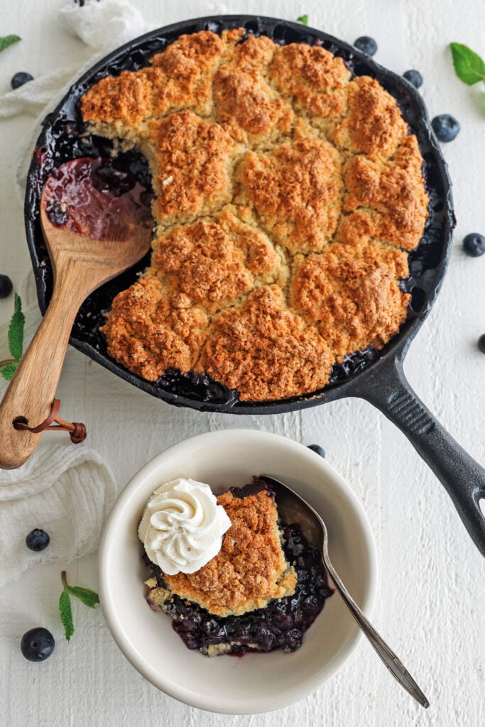 Blueberry cobbler in a skillet with a scoop in a bowl