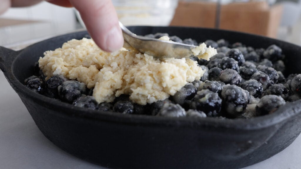 adding spoonfuls of batter to the blueberries in the skillet