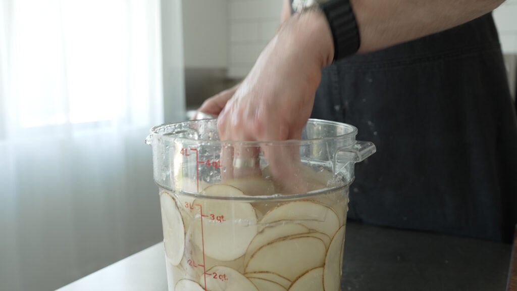 Thinly sliced potatoes in a large clear container filled with water and vinegar