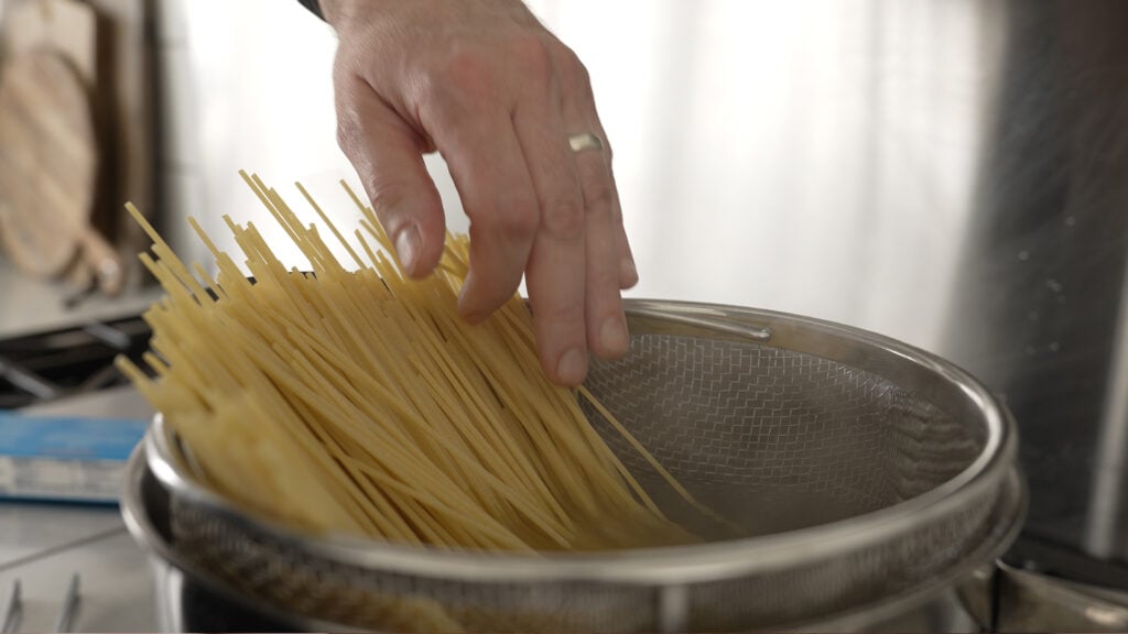 Adding spaghetti noodles to a large pot of water with a strainer inside