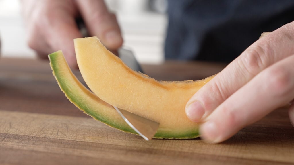 Using a knife to slice the rind from a wedge of melon