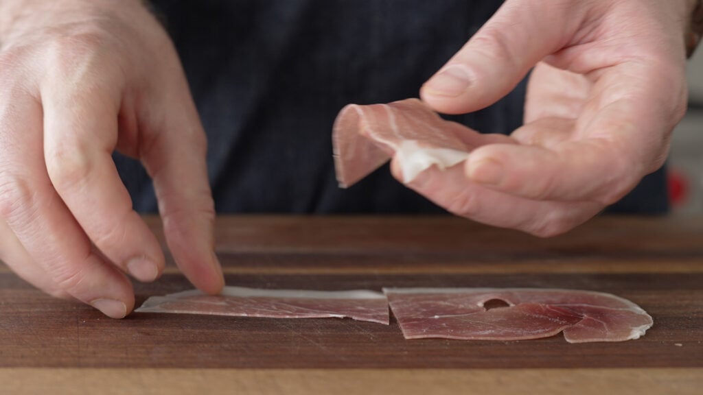 Picking up sliced prosciutto pieces from a cutting board