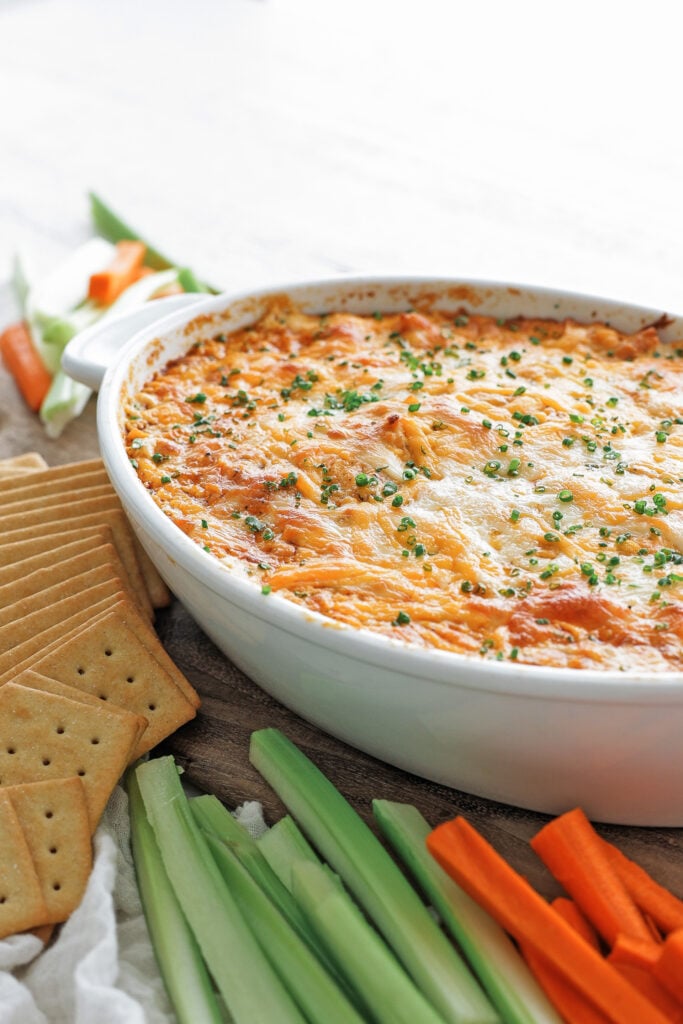Baked buffalo chicken dip in a white baking dish with a golden browned cheese top, served with celery, carrots, and tortilla chips