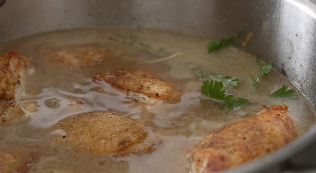 Seared chicken braising in stock with a tied herb bundle